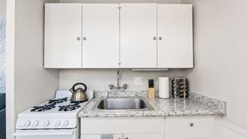 Une cuisine compacte avec des meubles blancs, un évier en acier inoxydable, une cuisinière à gaz et une bouilloire.