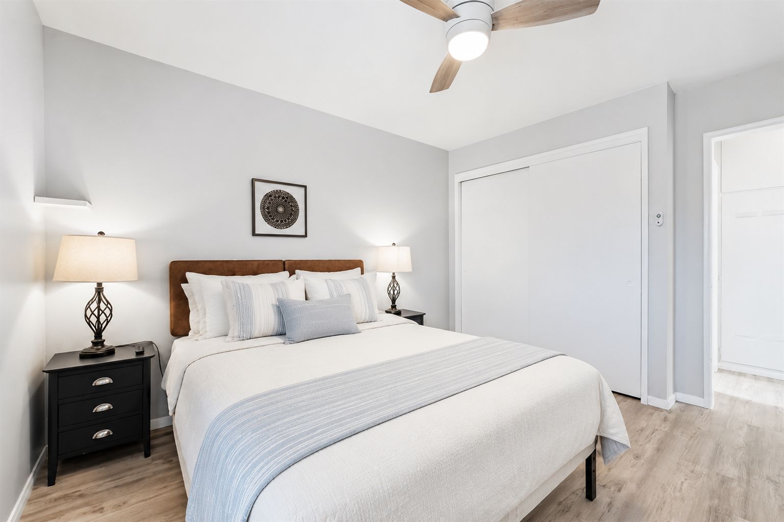 Une chambre à coucher comprenant un lit, des tables de chevet, un ventilateur de plafond, une porte et une œuvre d’art accrochée au mur.