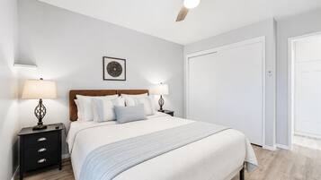 Une chambre à coucher comprenant un lit, des tables de chevet, un ventilateur de plafond, une porte et une œuvre d’art accrochée au mur.