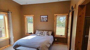 A bedroom with a bed, two windows, a bench, and a picture on the wall. - Peaceful Farmhouse with hot tub (Yankton)