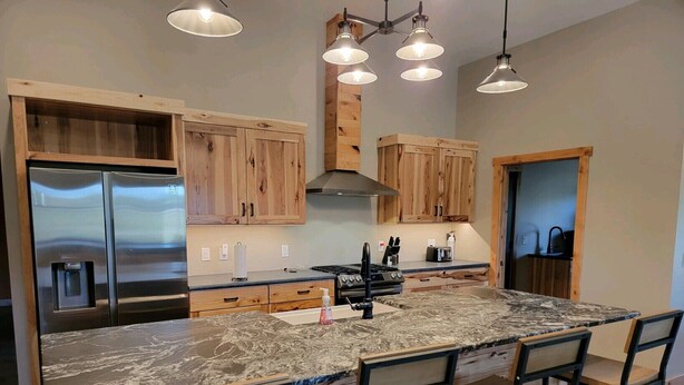 A modern kitchen with wooden cabinets, a stainless steel refrigerator, a marble island with bar stools, and pendant lighting. - Peaceful Farmhouse with hot tub (Yankton)