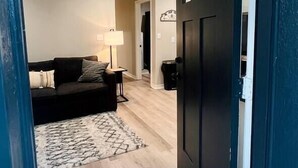 Una sala con un sofá de color oscuro, un tapete estampado, un ventilador de techo y una puerta que da a otra habitación.
