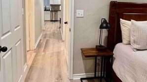 Un dormitorio con cabecera de madera, una mesita de noche con lámpara y vista a la cocina a través de una puerta abierta.