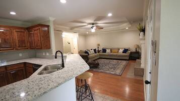 A kitchen with granite countertops and wooden cabinets, a dining area with a bar stool, and a living room with a sofa and a ceiling fan.