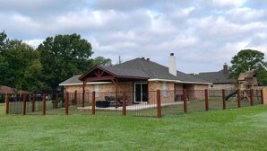A single-story brick building with a covered patio area, a fenced yard, and a playground structure in the background. - Royal Entertainer - STAYeasttexas (Chandler)