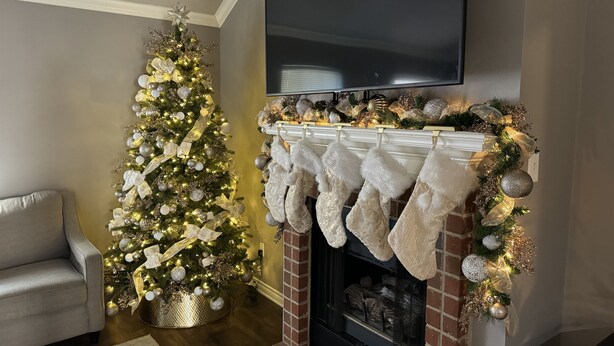 Un salon chaleureux avec un sapin de Noël décoré, une cheminée ornée de chaussettes et un téléviseur à écran plat fixé au-dessus.