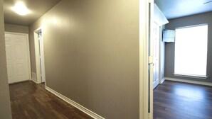 Un couloir avec un plancher en bois, une porte blanche et une fenêtre avec des stores.