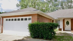 Une maison à deux étages avec une façade en briques, une porte de garage blanche et un jardin bien entretenu à l’avant, agrémenté d’une haie.