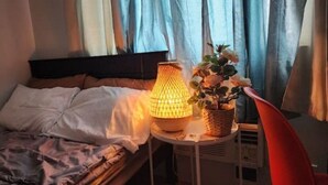 En säng med ett vitt överkast och kuddar, ett sängbord med en lampa, en korg med blommor och en röd stol.