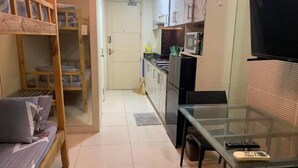 A compact kitchen with a microwave, stove, and cabinets. There is a glass table with a remote control and a small device on it. A bunk bed is visible in the background. - Sea Resi.MoA.Airport Netflix (Pasay)