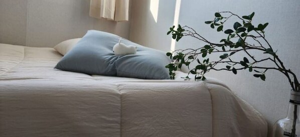 A neatly made bed with a light blue pillow and a white pillow, a vase with a branch of green leaves, and a window with curtains.