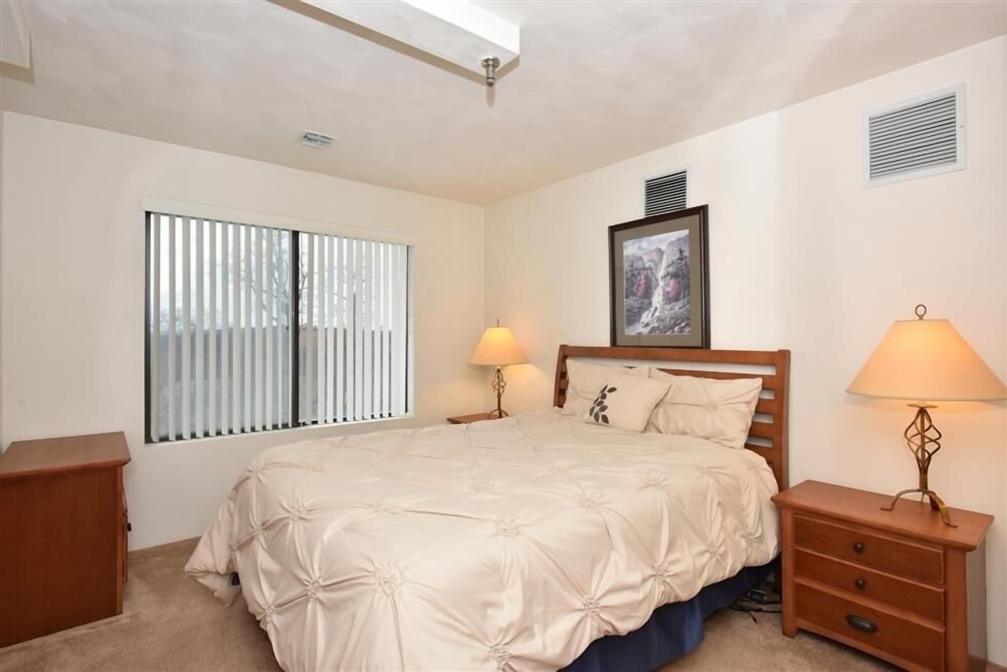 A bedroom with a large bed, two bedside tables with lamps, a window with blinds, a framed picture on the wall, and a wooden nightstand.