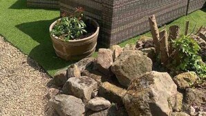 Un edificio in pietra con tetto a falda, pannelli solari e un’area giardino con un divano in vimini, un vaso grande e rocce.