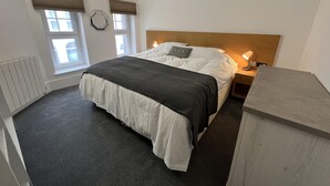 A bedroom with a bed, bedside tables, a headboard, a radiator, and two windows with blinds. - Charming 1-bed & Balcony Flat in Merthyr Tydfil (Merthyr Tydfil)