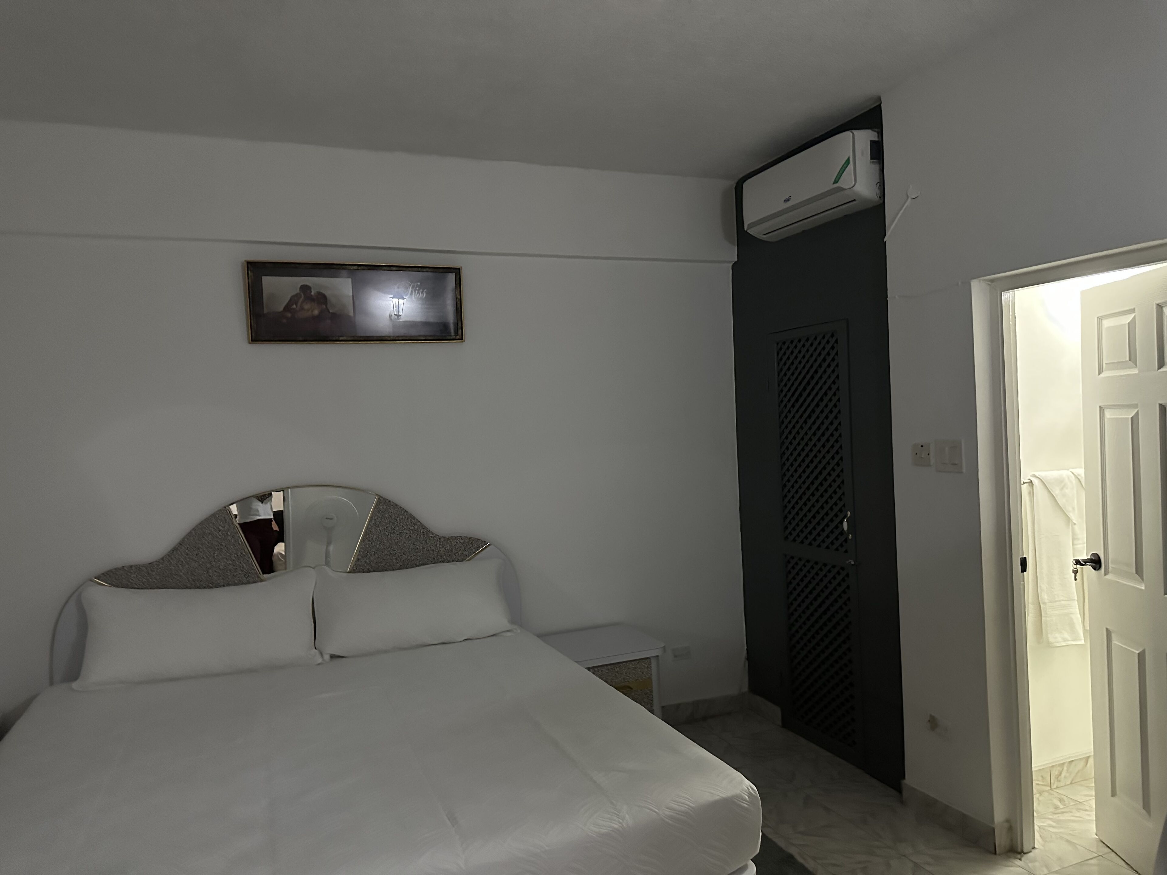 Un dormitorio con cama, aire acondicionado instalado en la pared, una puerta que da a otra habitación y un cuadro en la pared.