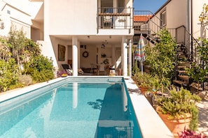 Piscina com deck de azulejos, rodeada por plantas em vasos e área de estar externa com mobiliário.