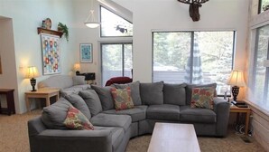 A living room with a grey sectional sofa, a wooden coffee table, and two side tables with lamps. There are large windows with blinds, a chandelier, and artwork on the walls. - Seven Springs 3 BR Condo, Loft and Mountain Views! by RedAwning (Champion)