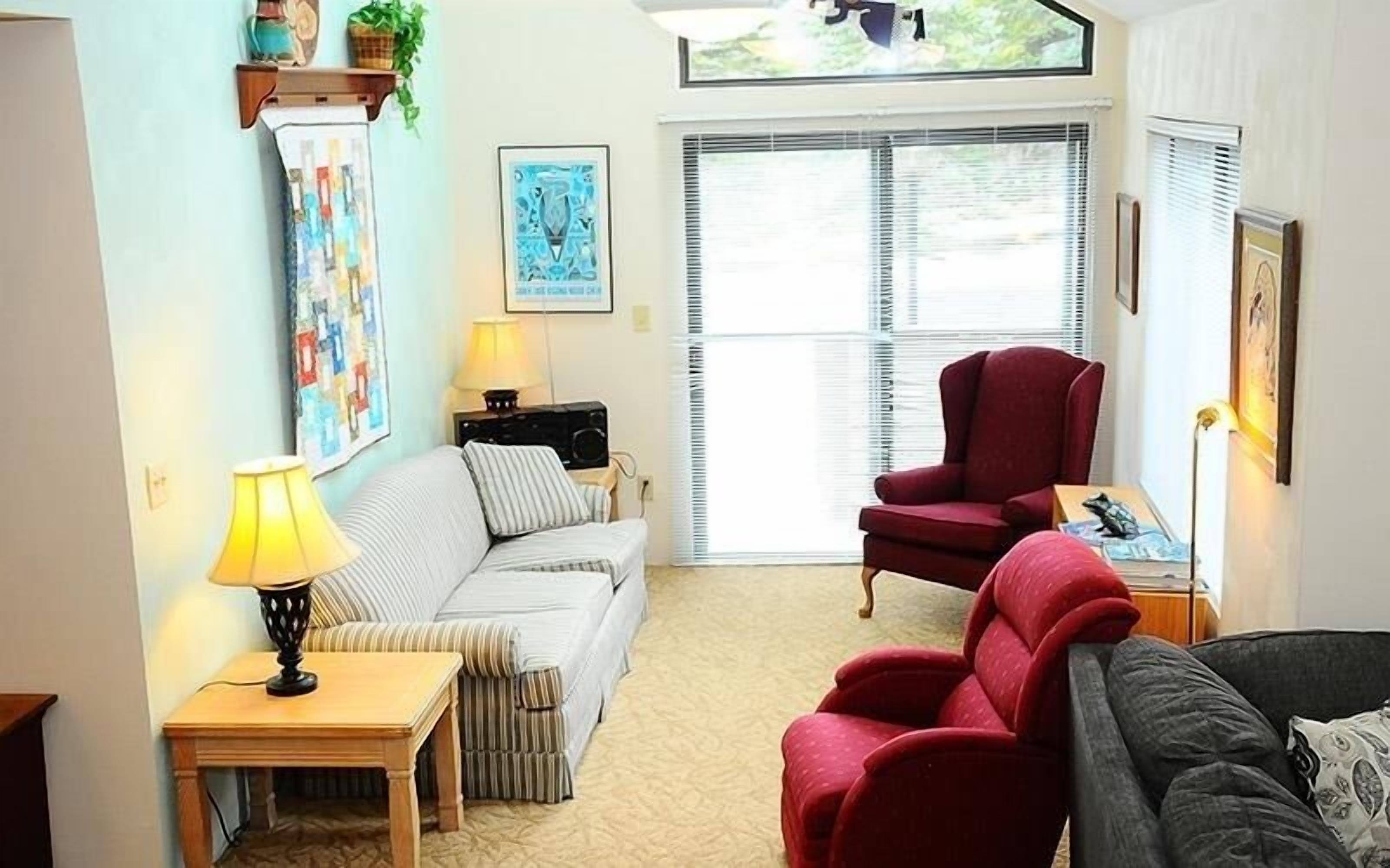A living room with a sofa, armchairs, a coffee table, and a lamp. There are two windows with blinds, a painting on the wall, and a small table with a lamp.