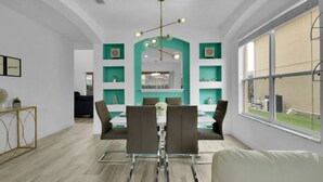 A modern dining area with a central table, chairs, and a chandelier. There are teal shelves on the walls and a large window on the right.