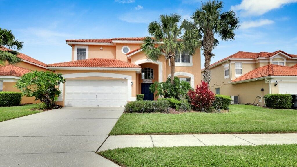 A two-story house with a red-tiled roof, a garage, and a well-maintained lawn with palm trees and shrubs.