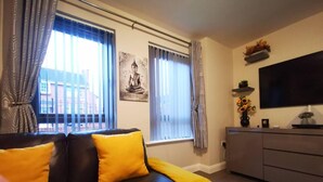 Un salon avec un canapé en cuir noir, deux coussins jaunes, une statue de Bouddha, un téléviseur à écran plat et une table basse en verre.
