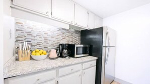 Una cocina con armarios blancos, salpicadero de azulejos, refrigerador de acero inoxidable, microondas, cafetera y un bol con limones.