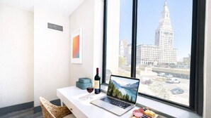 Un espacio de trabajo moderno con una computadora portátil, vino y aperitivos junto a un amplio ventanal con vistas a un paisaje urbano.
