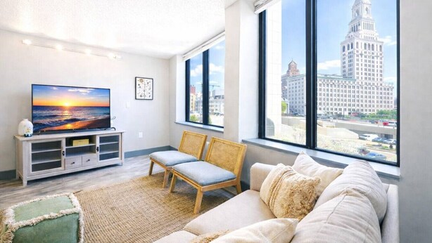 Una sala de estar moderna con un televisor grande, un sofá y una silla, con vistas a la ciudad a través de amplios ventanales.