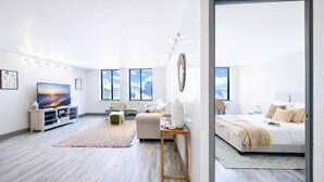 Amplia sala de estar con un televisor grande, un sofá, una mesa de centro y una alfombra. Varias ventanas ofrecen vistas a la ciudad.