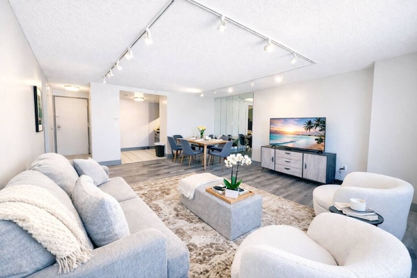A modern living room with a grey sofa, white armchairs, a coffee table, and a TV on a wooden cabinet.