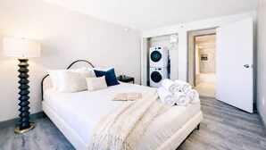 Moderni hotellihuone, jossa on suuri sänky, yöpöytälamppu, kirja ja pyyhesetti. Taustalla näkyy pesukone ja kylpyhuone.