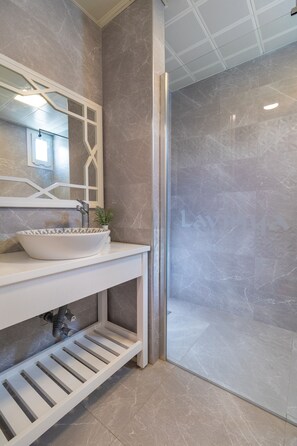 Een badkamer met een wit wastafelmeubel, een wasbak in een kom, een spiegel met een geometrisch kader, een kleine potplant en een douche met een glazen deur.