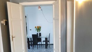 Un couloir mène à une salle à manger où se trouvent une table et des chaises, un vase rempli de tournesols et un luminaire fixé au mur.