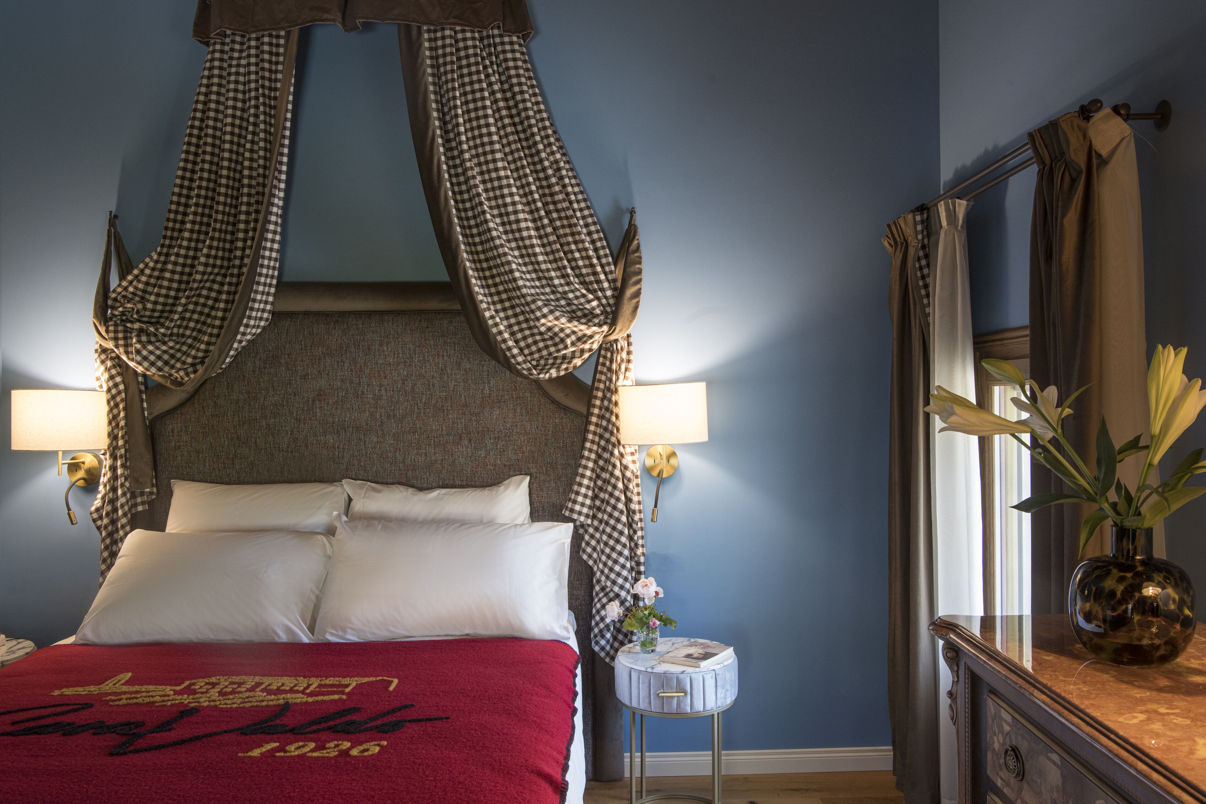 A bedroom with a bed, pillows, a bedside table, a lamp, a mirror, and a vase with flowers.