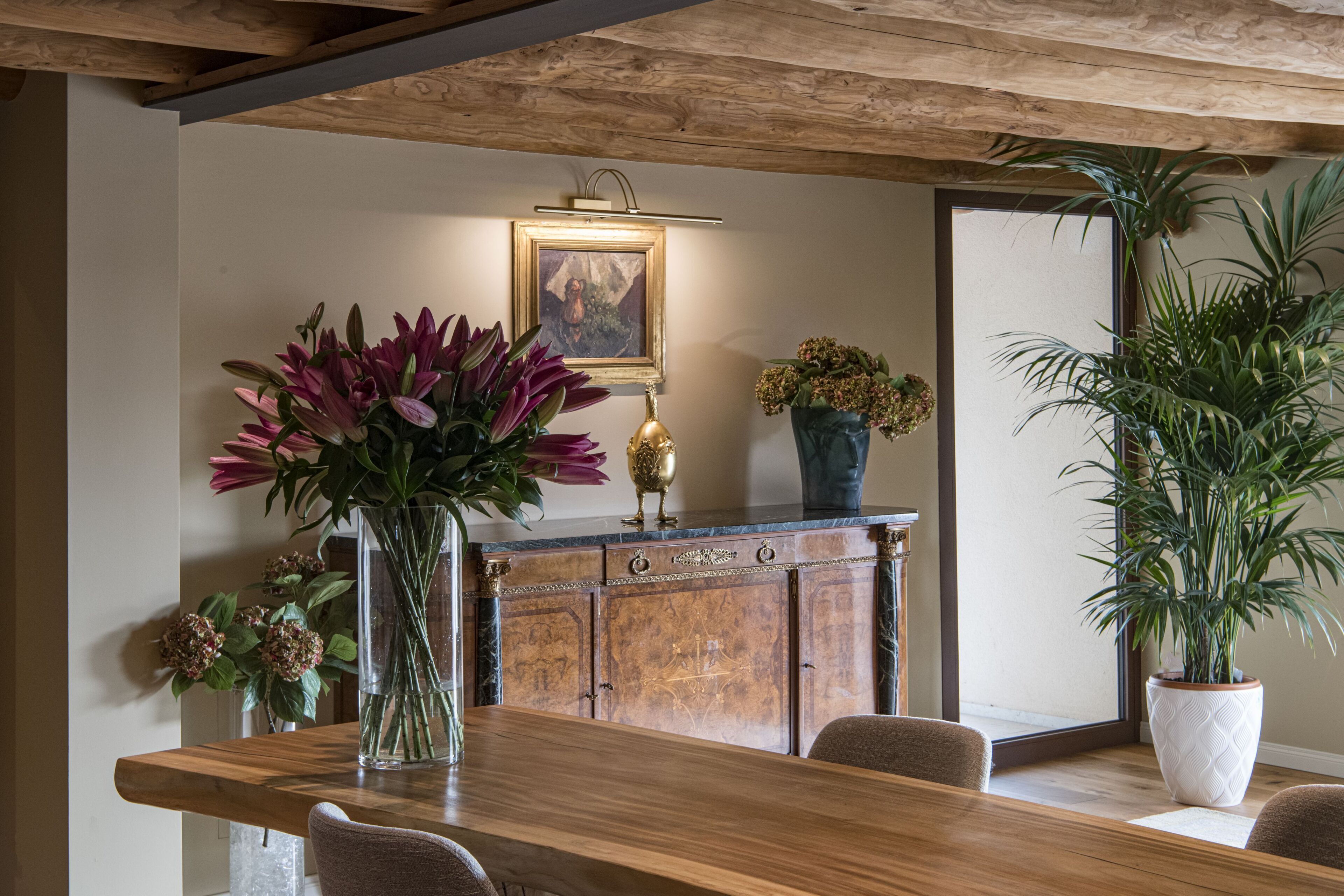 Un comedor con mesa de madera, sillas, un jarrón de flores, un aparador con un cuadro y una planta en maceta.
