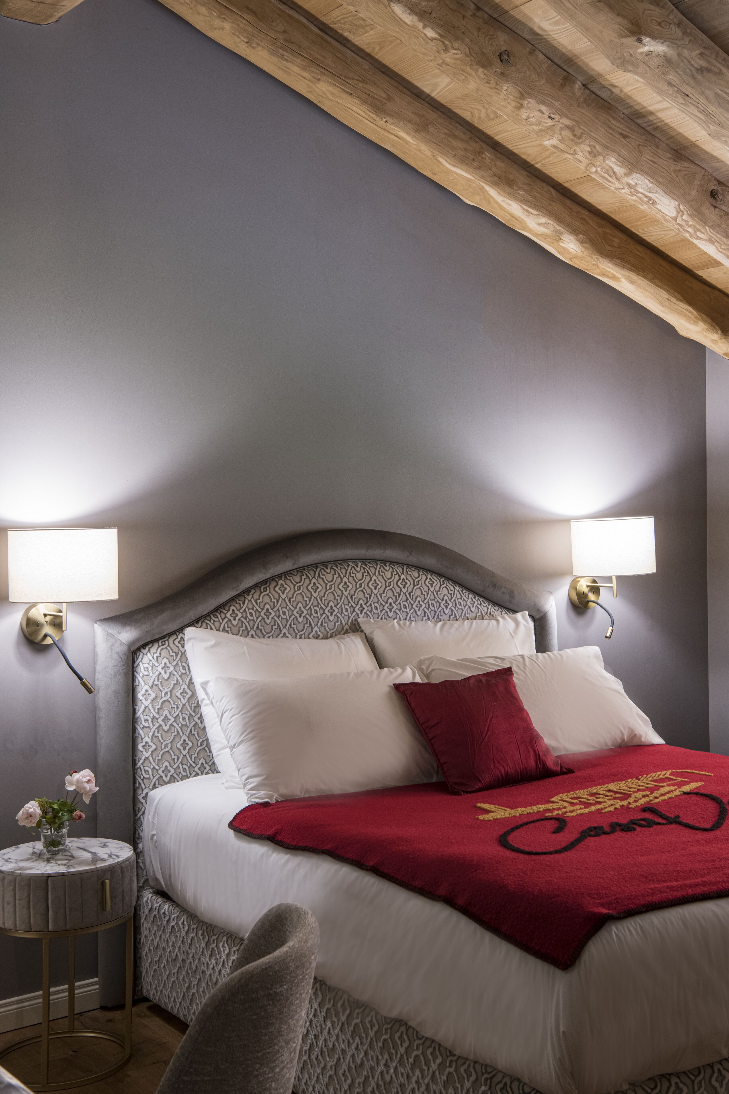 A bedroom with a bed, two bedside lamps, a round table with a vase of flowers, and a red throw blanket.