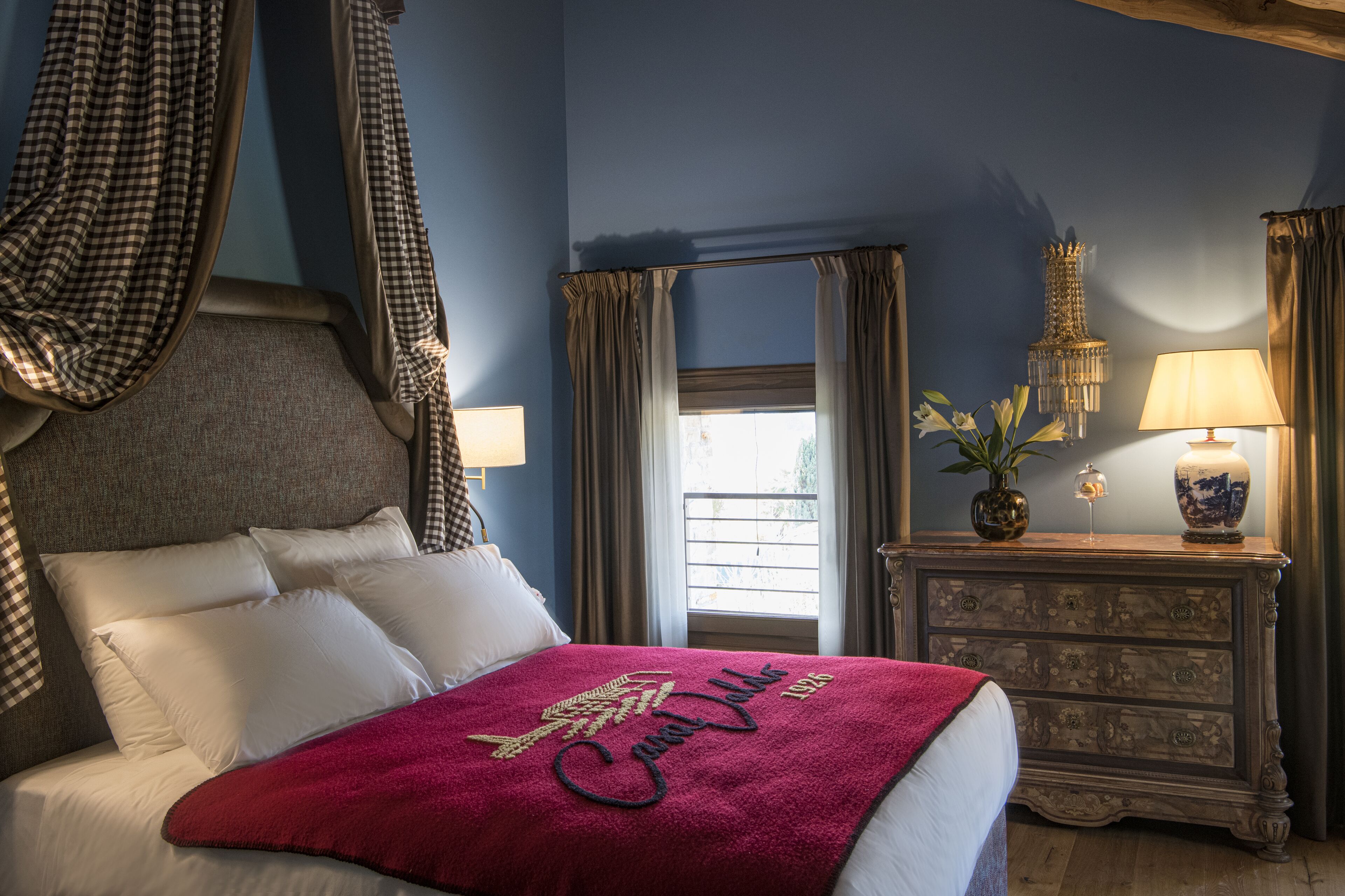 A bedroom with a bed, a nightstand, a dresser, a window with curtains, and a chandelier.