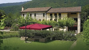 Un bâtiment à deux étages, doté d’une façade en pierre et d’un toit en tuiles, entouré d’une végétation luxuriante et avec une chaîne de montagnes en arrière-plan.