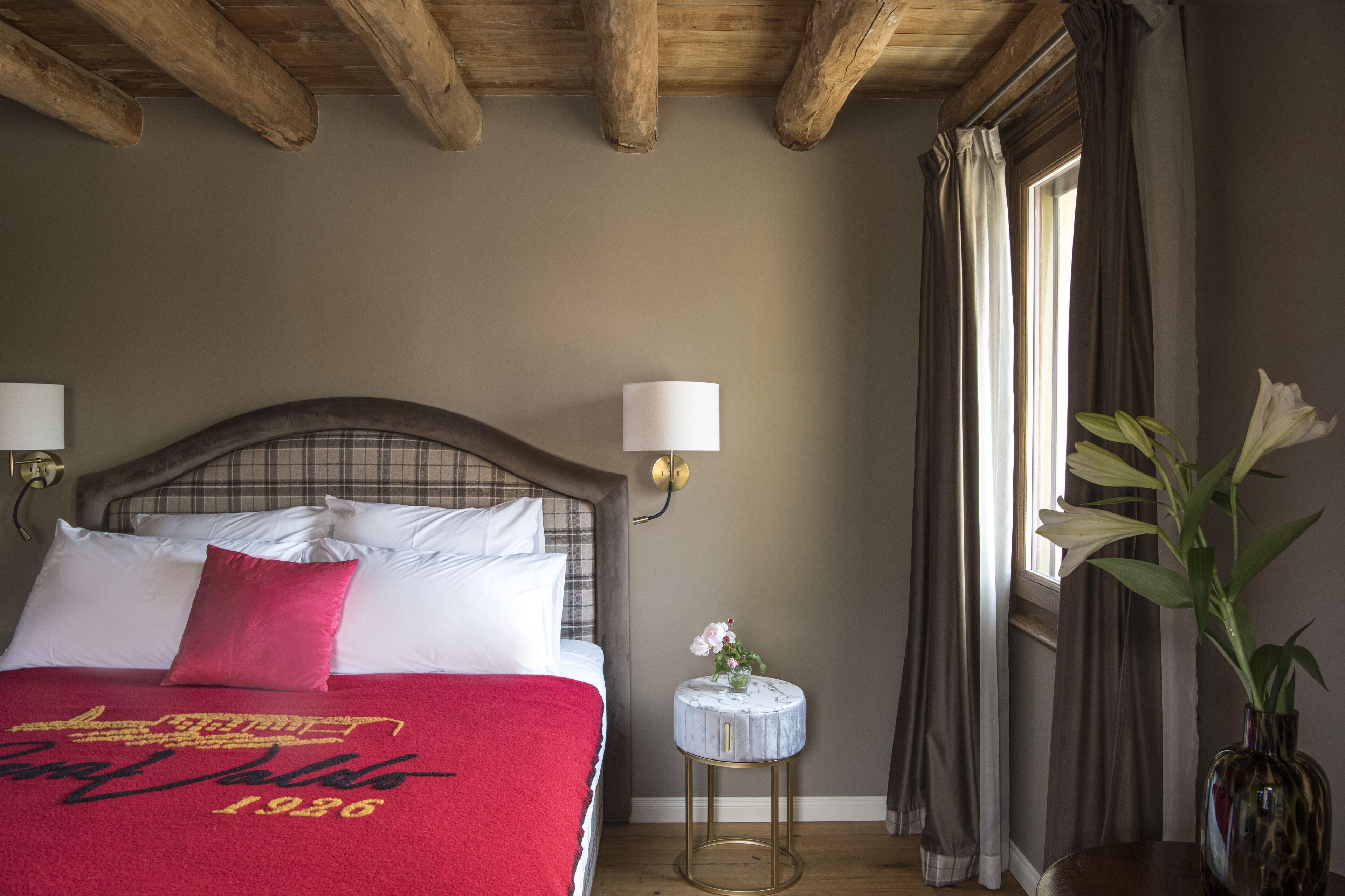 A bedroom with a bed, pillows, a bedside table with a vase, and a window with curtains.