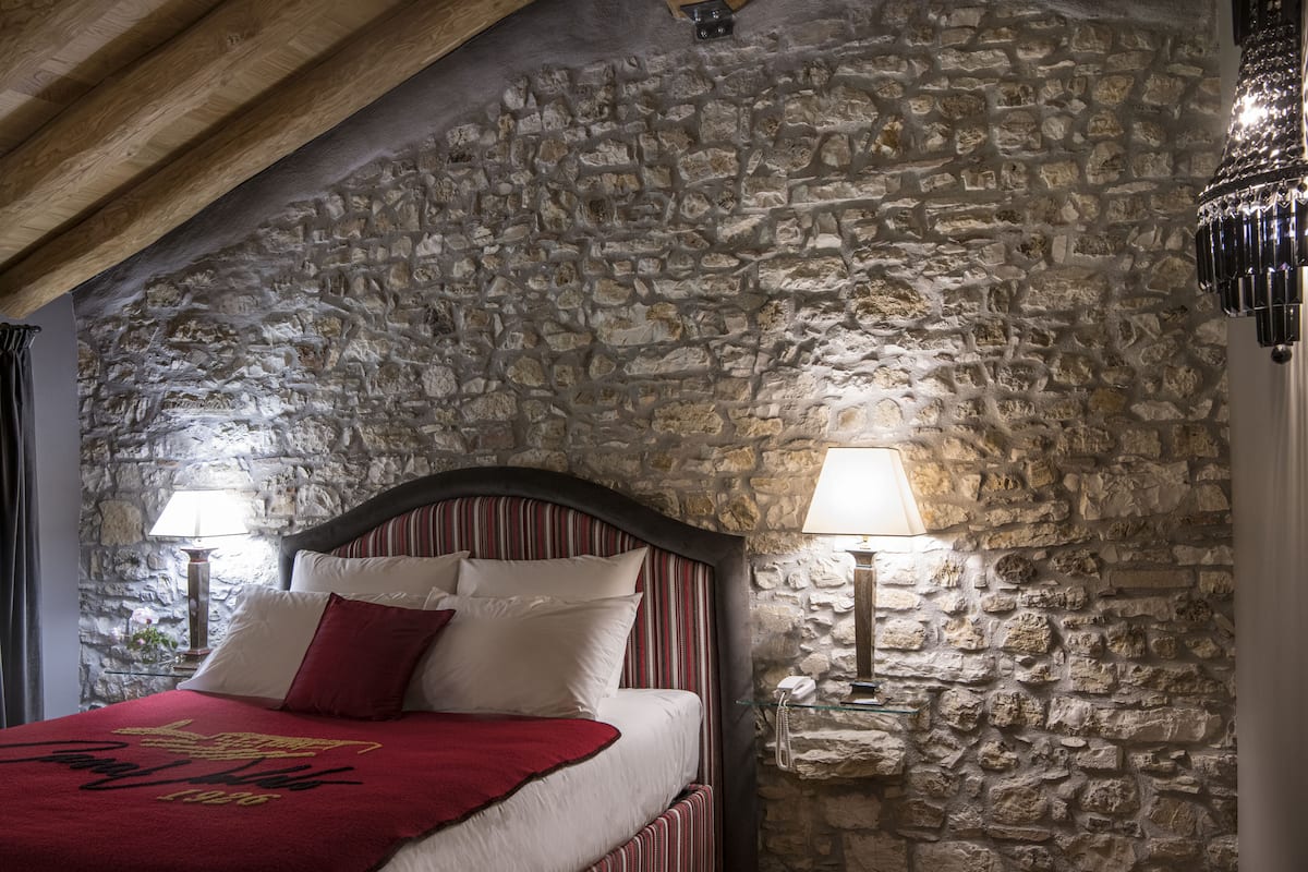 Ein Schlafzimmer mit einer Steinsandsteinwand, einer Holzbalkendecke, einem Bett mit weißer und roter Bettwäsche sowie zwei Nachttischlampen.