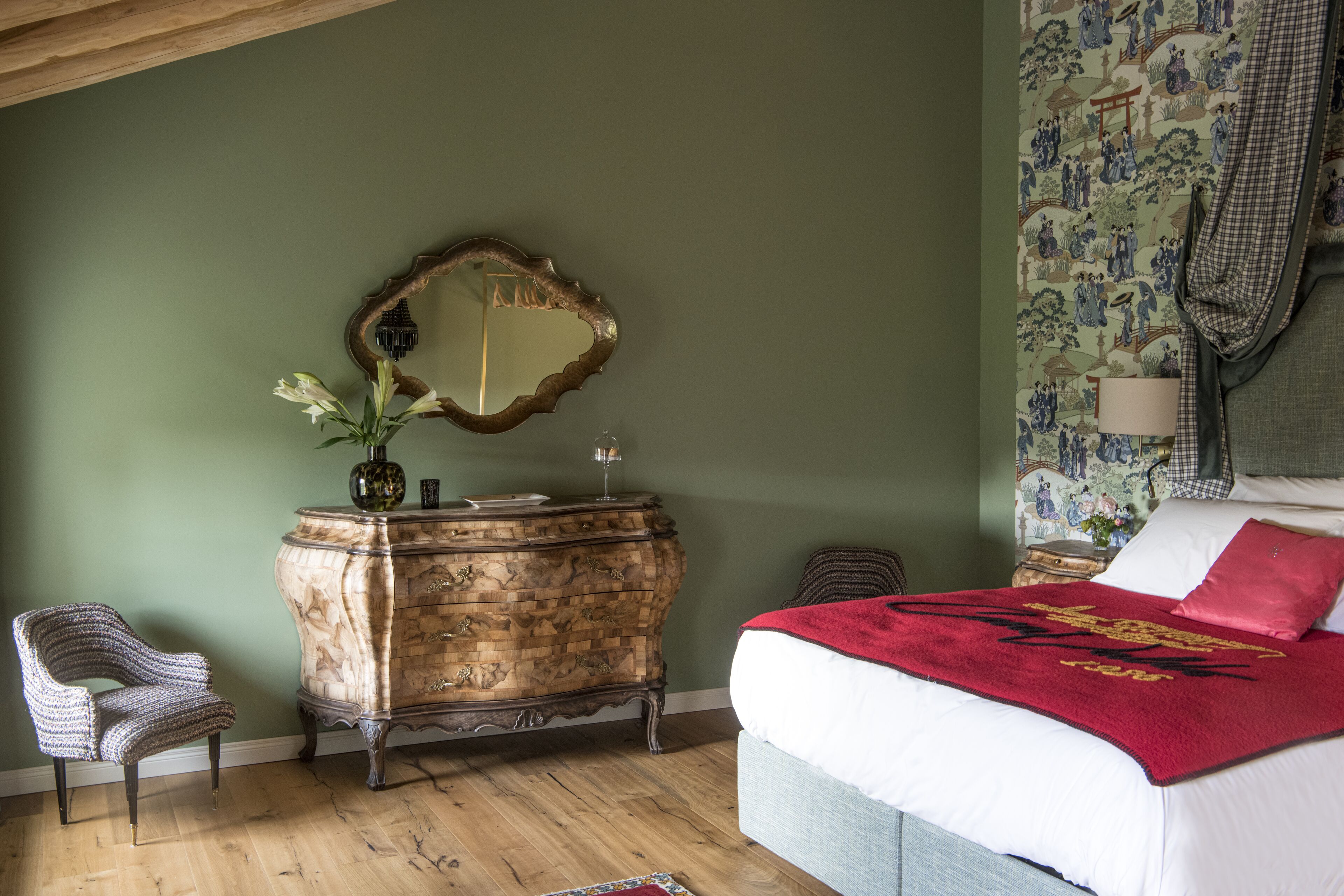 A bedroom with a wooden dresser, a mirror, a bed with a red blanket, and a chair.