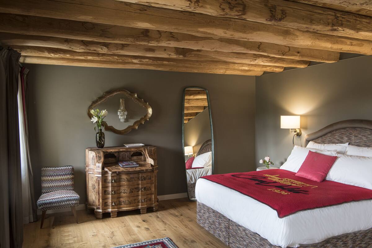 Ein Schlafzimmer mit Holzbalkendecke, einem Bett mit weißer und roter Bettwäsche, einem Schminktisch, einem Stuhl und einem Spiegel.