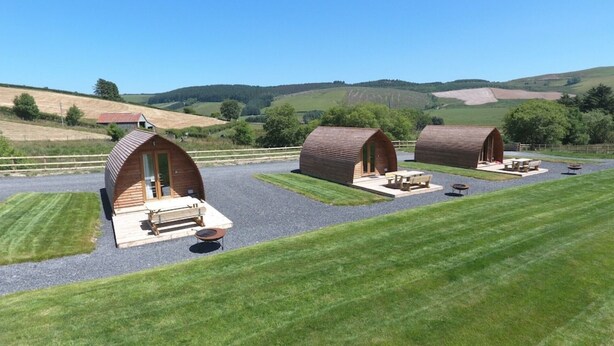 Entorno rural con tres cabañas de madera, cada una con techo curvo y un pequeño porche. Las cabañas están rodeadas de céspedes cuidados y senderos de grava.