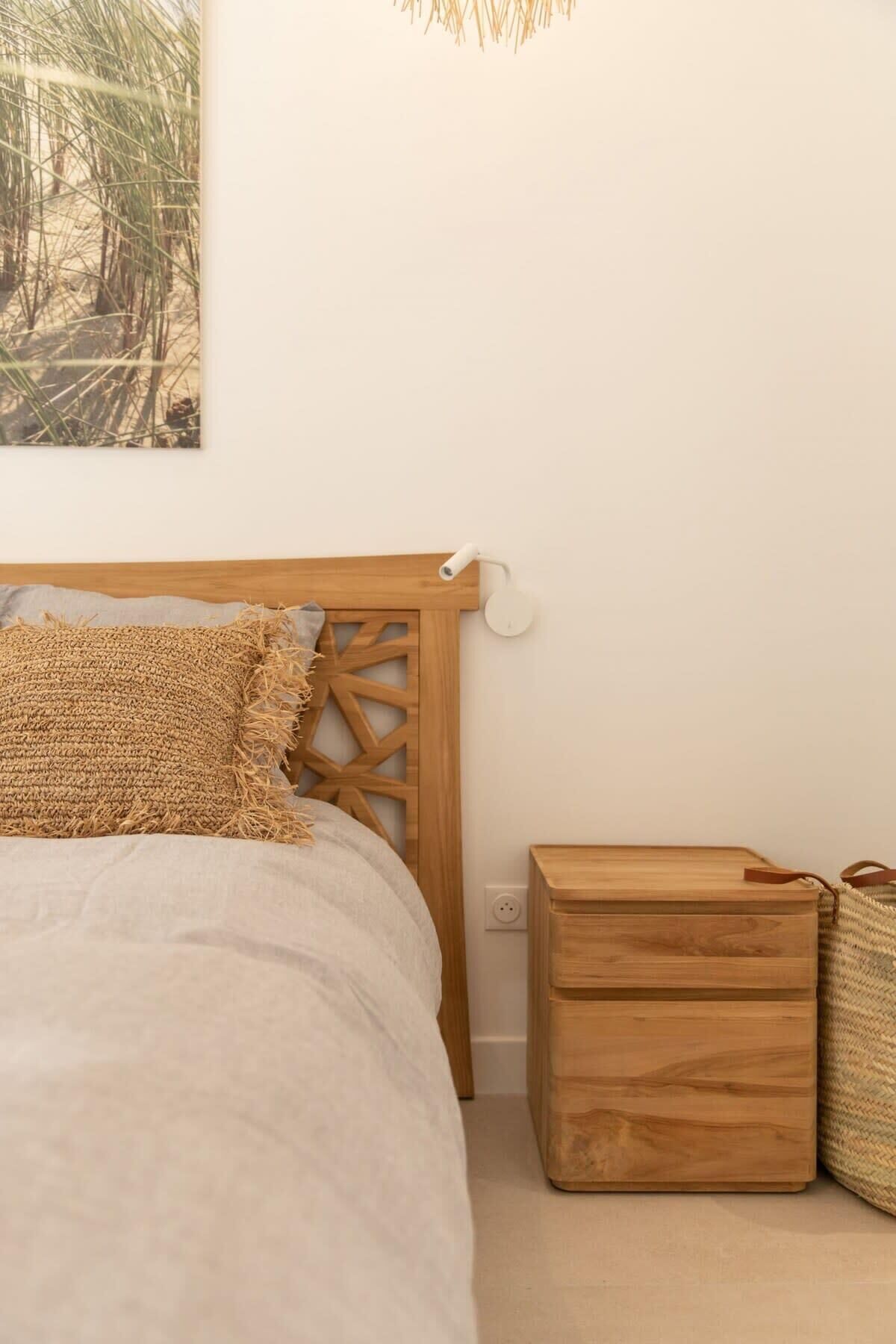 Une chambre à coucher avec une tête de lit en bois, un lit recouvert d’une couverture à motifs, une table de chevet surmontée d’un panier en osier et une lampe fixée au mur.