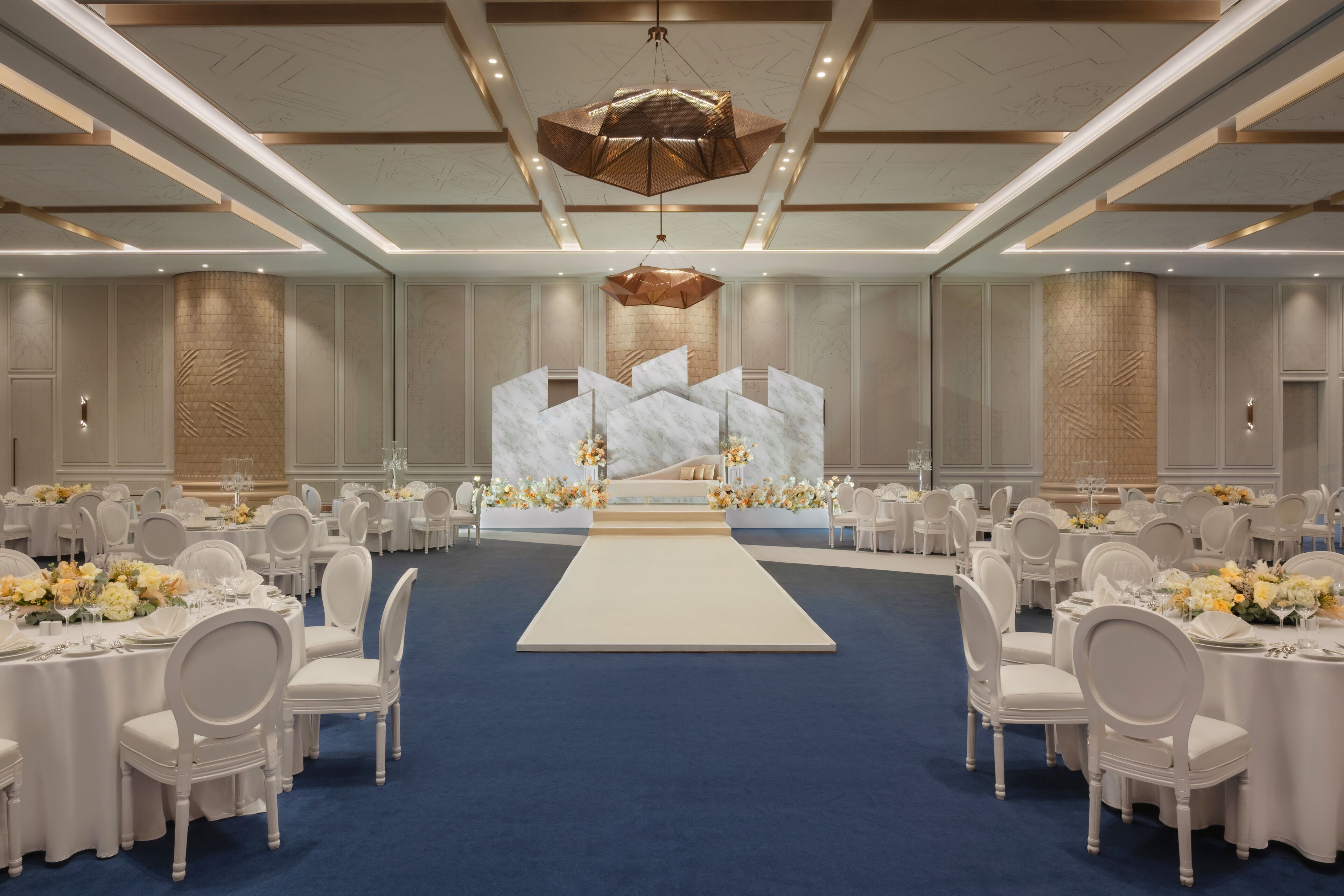 A formal event space with round tables, white chairs, a central stage, and a blue carpet.