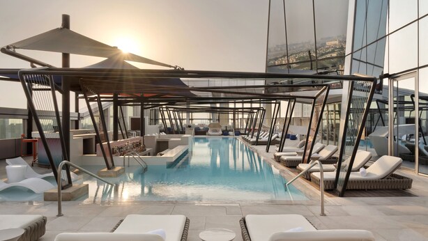 A rooftop pool with sun loungers and a covered seating area, surrounded by modern architecture.