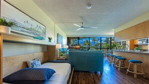 Ett modernt hotellrum med en säng, en soffa, en bardisk med barstolar och ett stort fönster med utsikt över palmer.