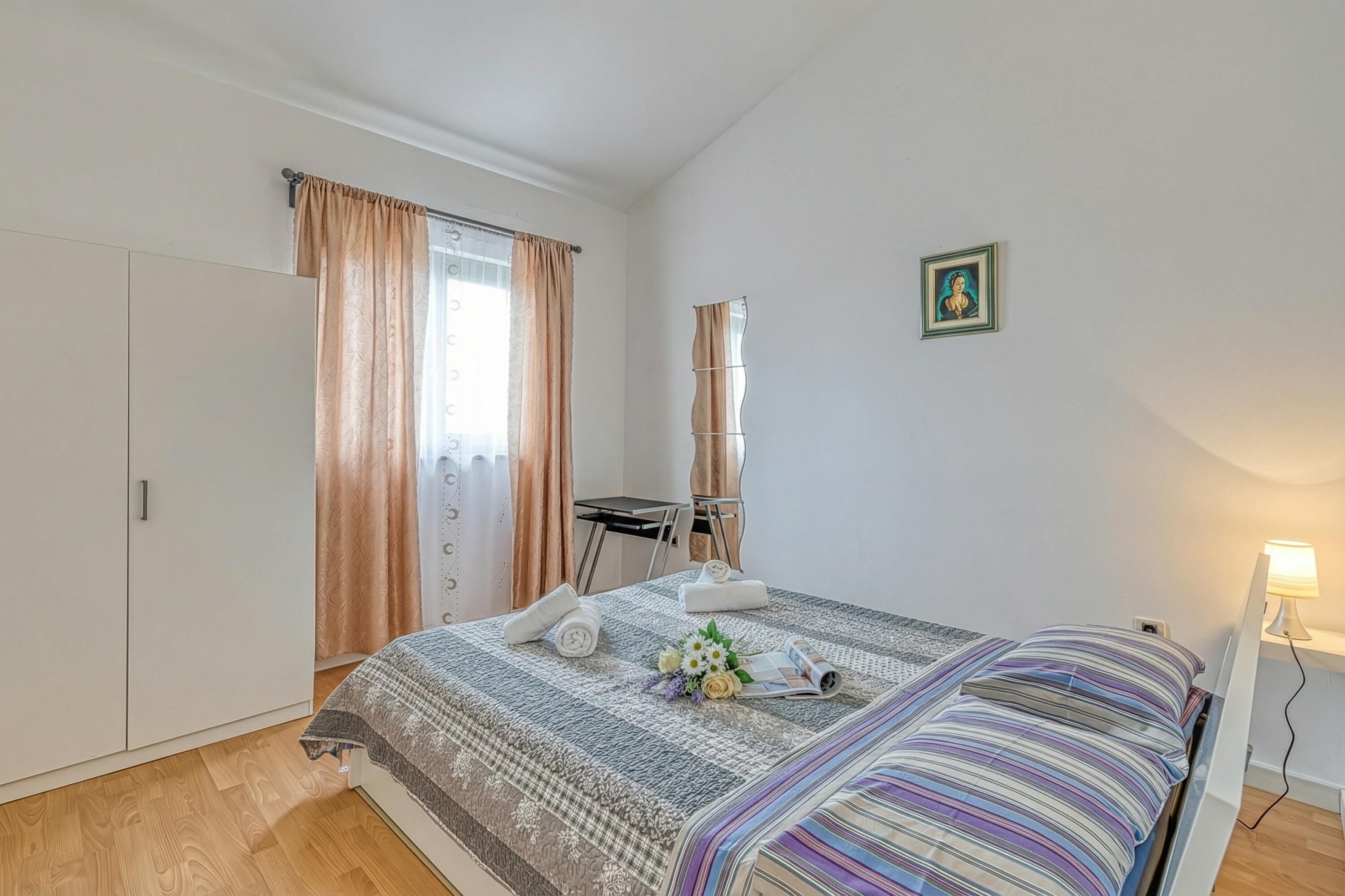 A bedroom with a bed, a desk, a chair, a small table, a picture on the wall, and a lamp.