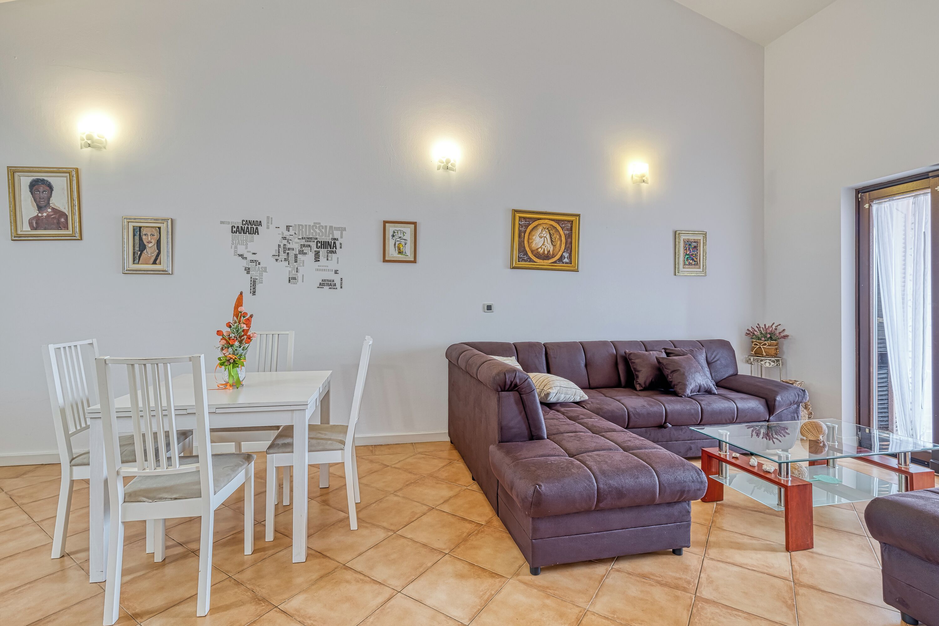 A living room with a brown sofa, white chairs, a glass coffee table, and a dining table with chairs. There are wall-mounted lights and framed pictures on the wall.