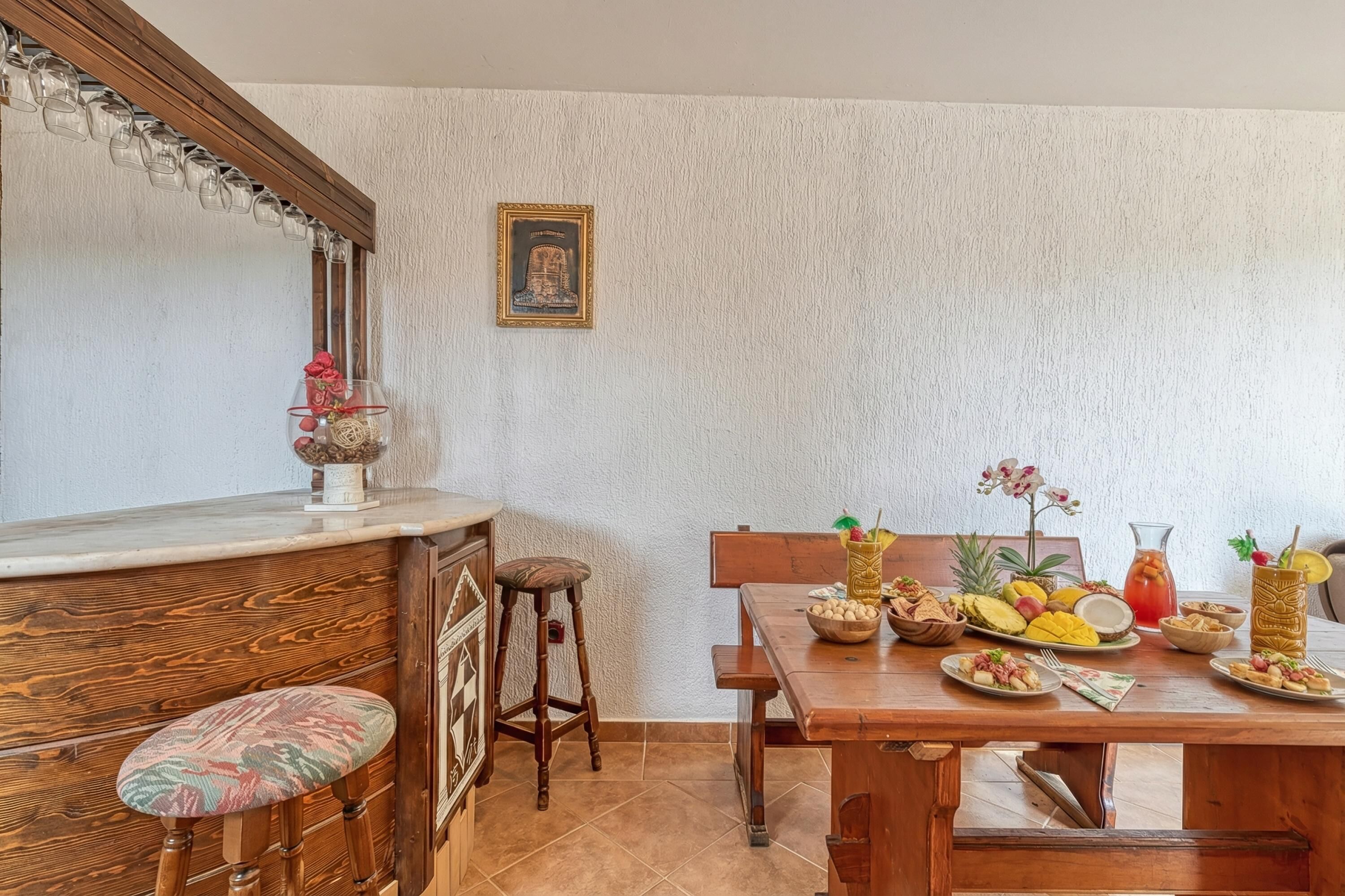 Un comedor con una mesa de madera dispuesta para una comida, una zona de bar con taburetes y un cuadro colgado en la pared.